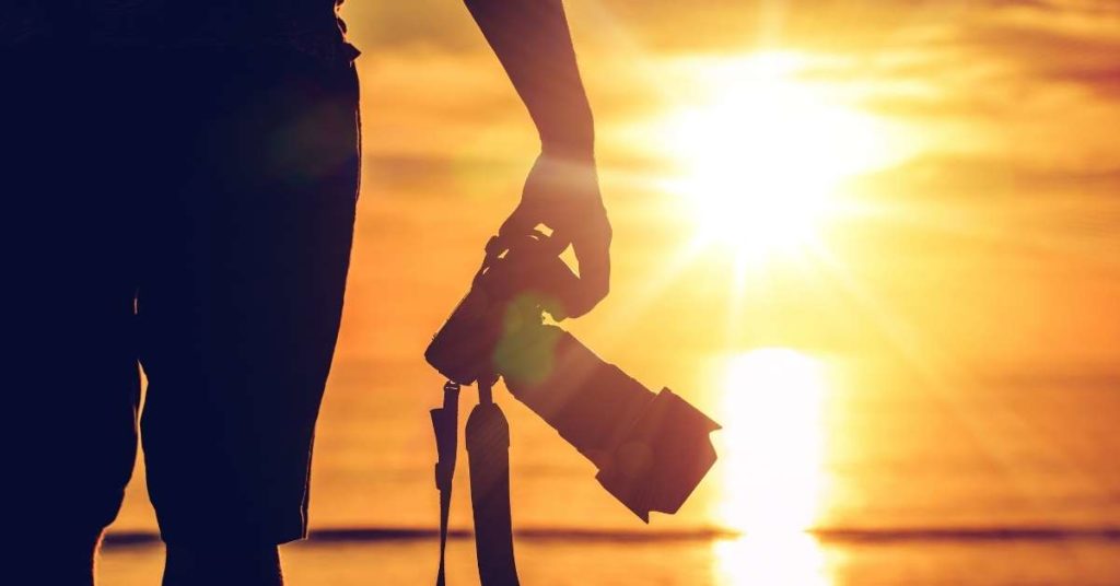 sombra das pernas de um fotógrafo em primeiro plano, ele segura uma câmera profissional em mãos. Ao fundo, cenário de pôr do sol na praia.