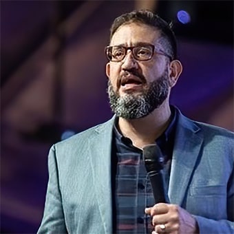 Homem de meia idade com blazer palestrando em salão de eventos.